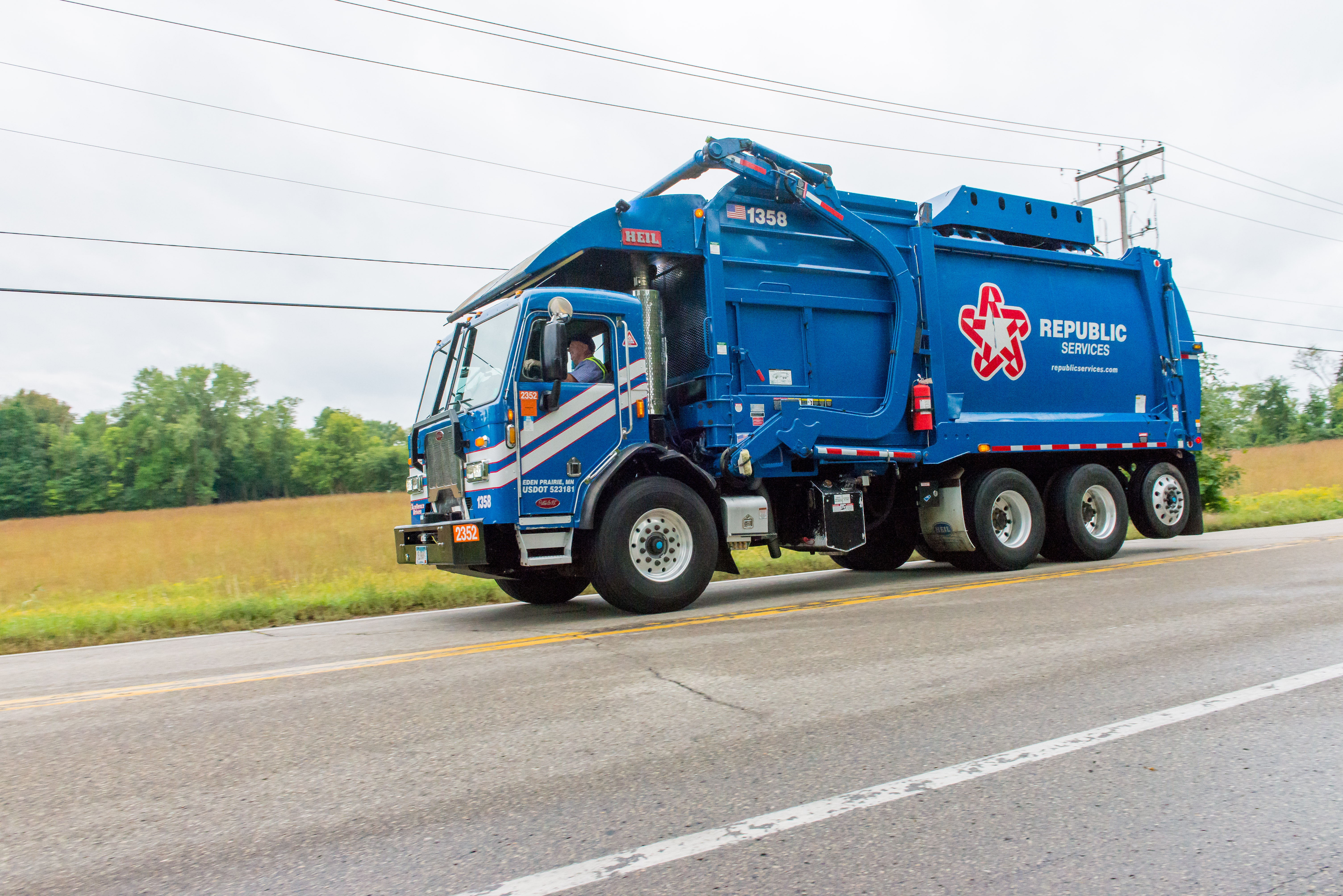 Blue Republic Services waste collection truck drives along street with grass and trees in the background.